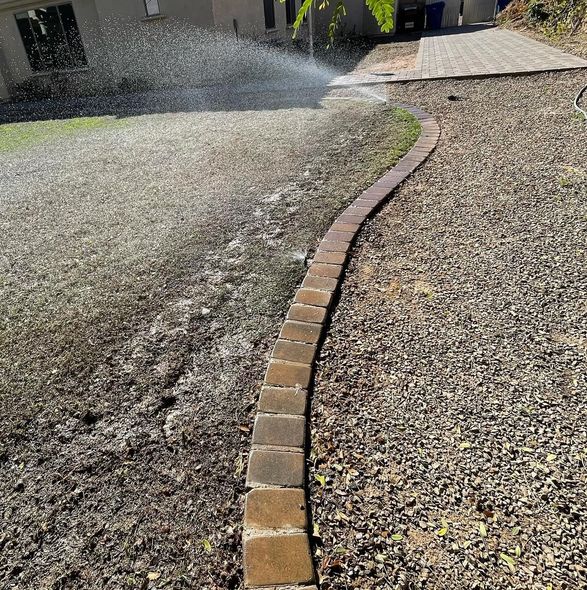 Sprinkler spraying water on lawn and gravel path lined with brick pavers.