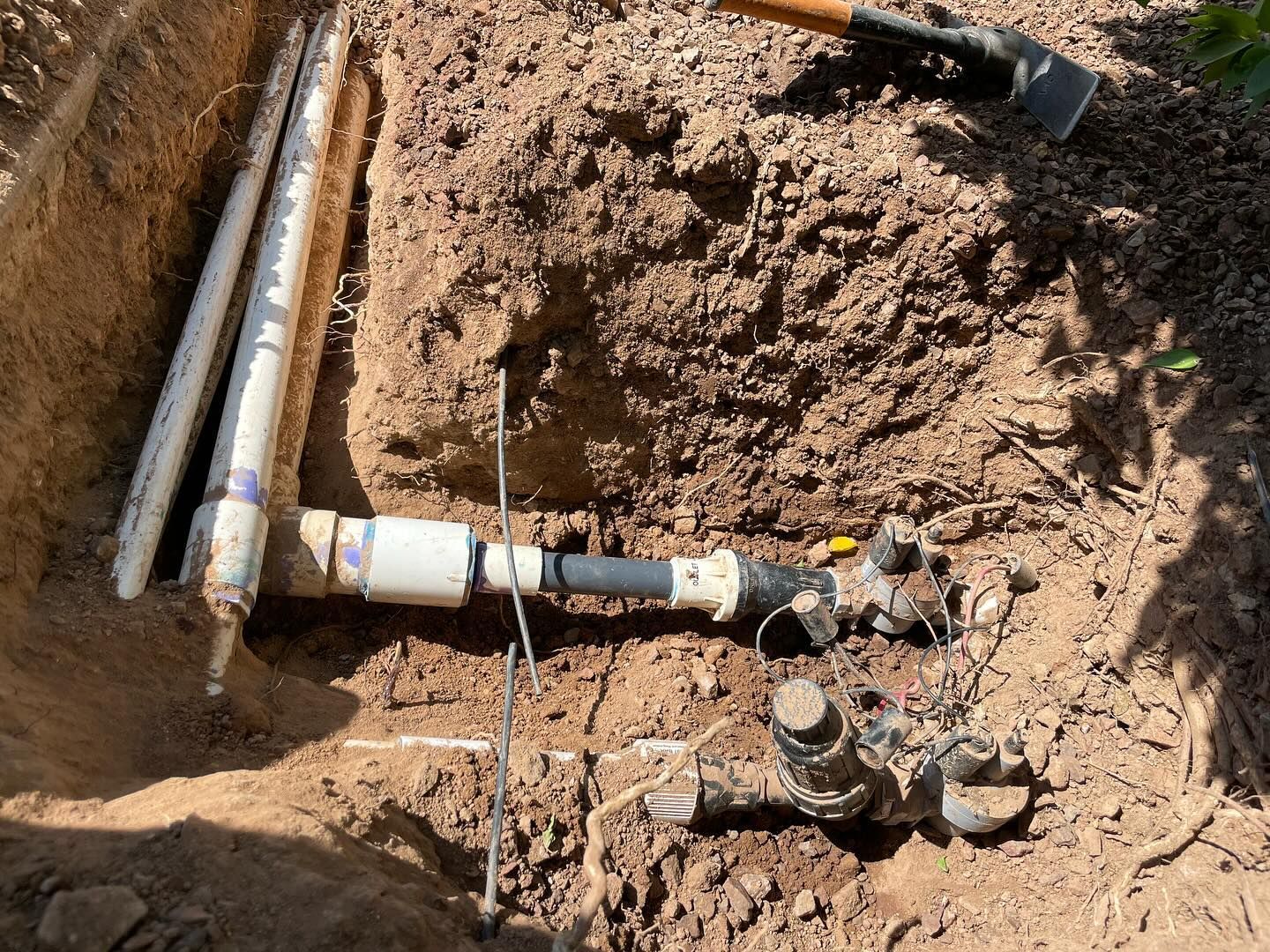 A dug-out trench showing exposed sprinkler system pipes, with dirt, a hammer, and white and black plastic pipes.