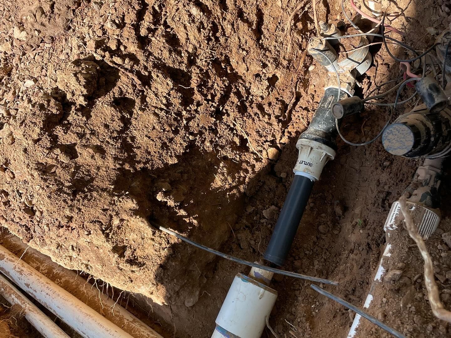 A buried irrigation sprinkler head with black pipe, white PVC, and surrounding brown soil.