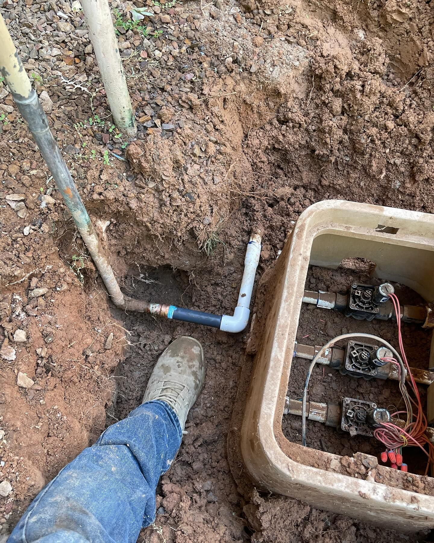 A person’s foot near an irrigation valve box with connected pipes in a dirt trench.