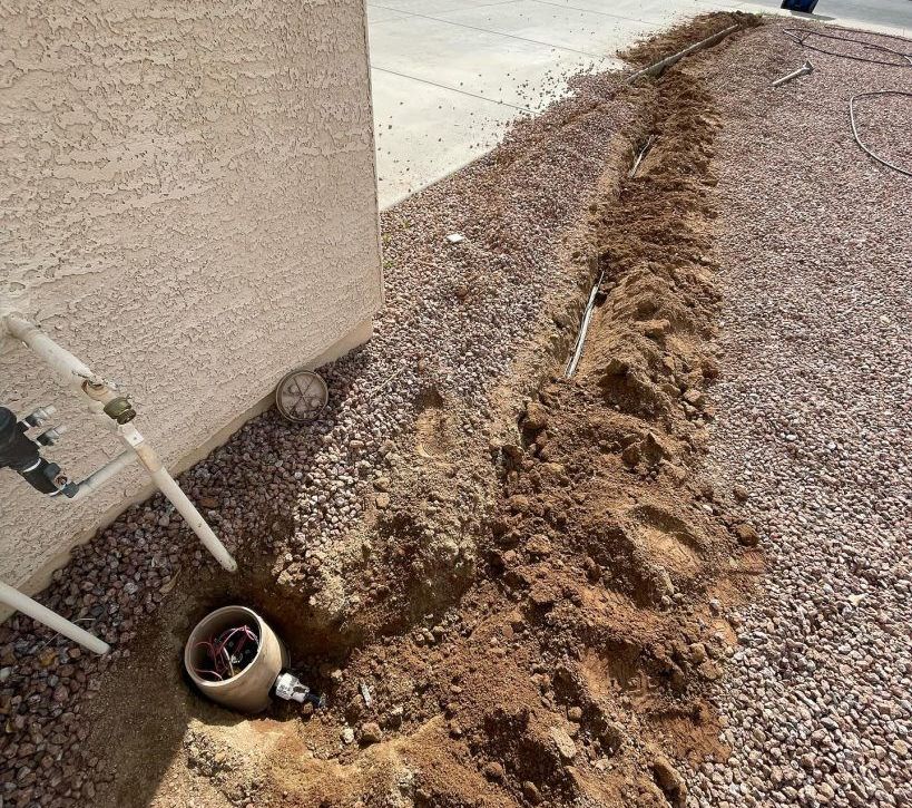 A trench dug in gravel next to a building, with pipes and dirt visible.