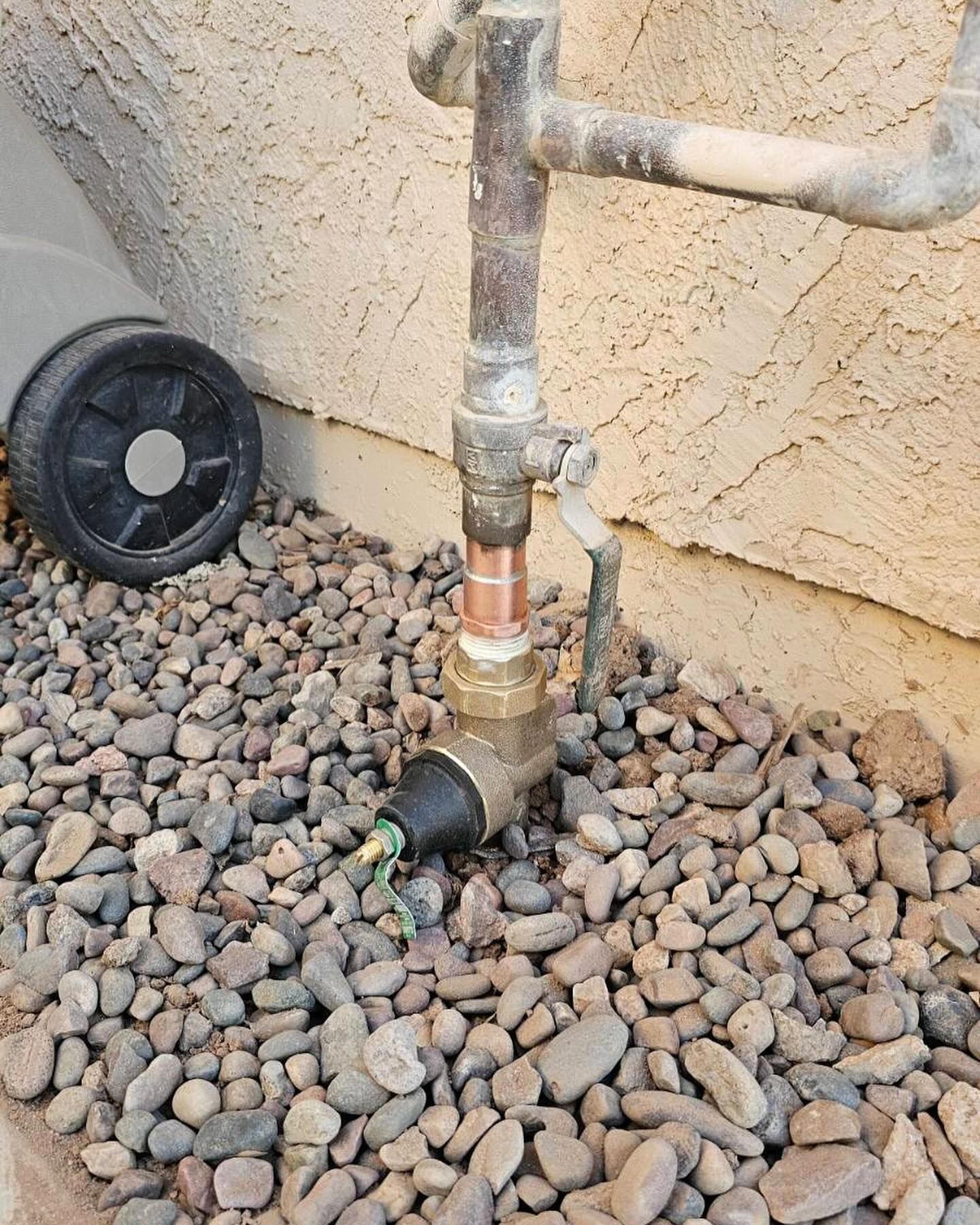 Outdoor plumbing with pipes, valves, and wiring amidst decorative gravel near a stucco wall.