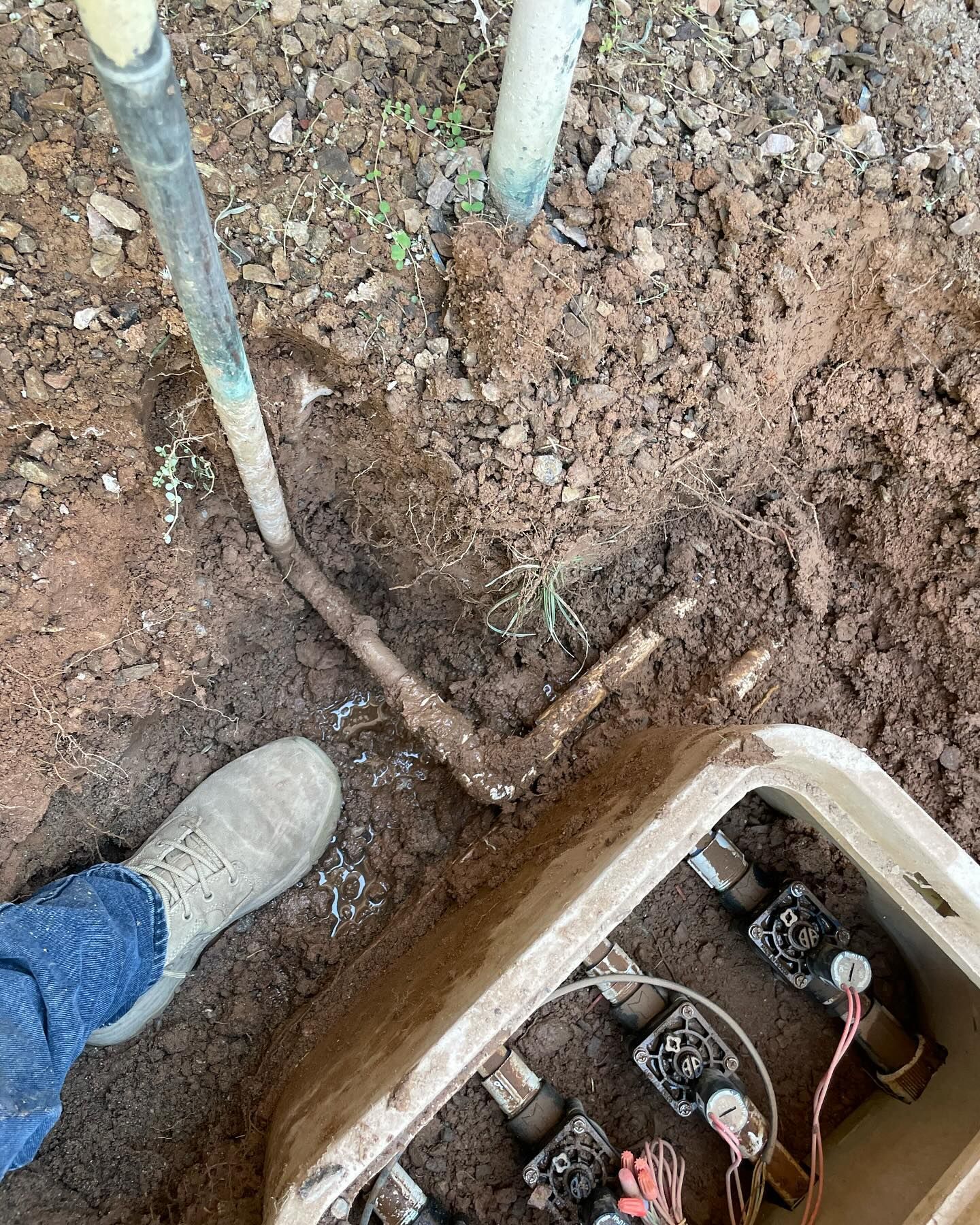 Person's foot and shovel near a brown irrigation box. A pipe is exposed in the dirt.