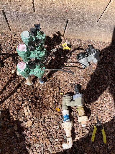 Irrigation system components: green valves, pipes, and a pump on gravel, outdoors.