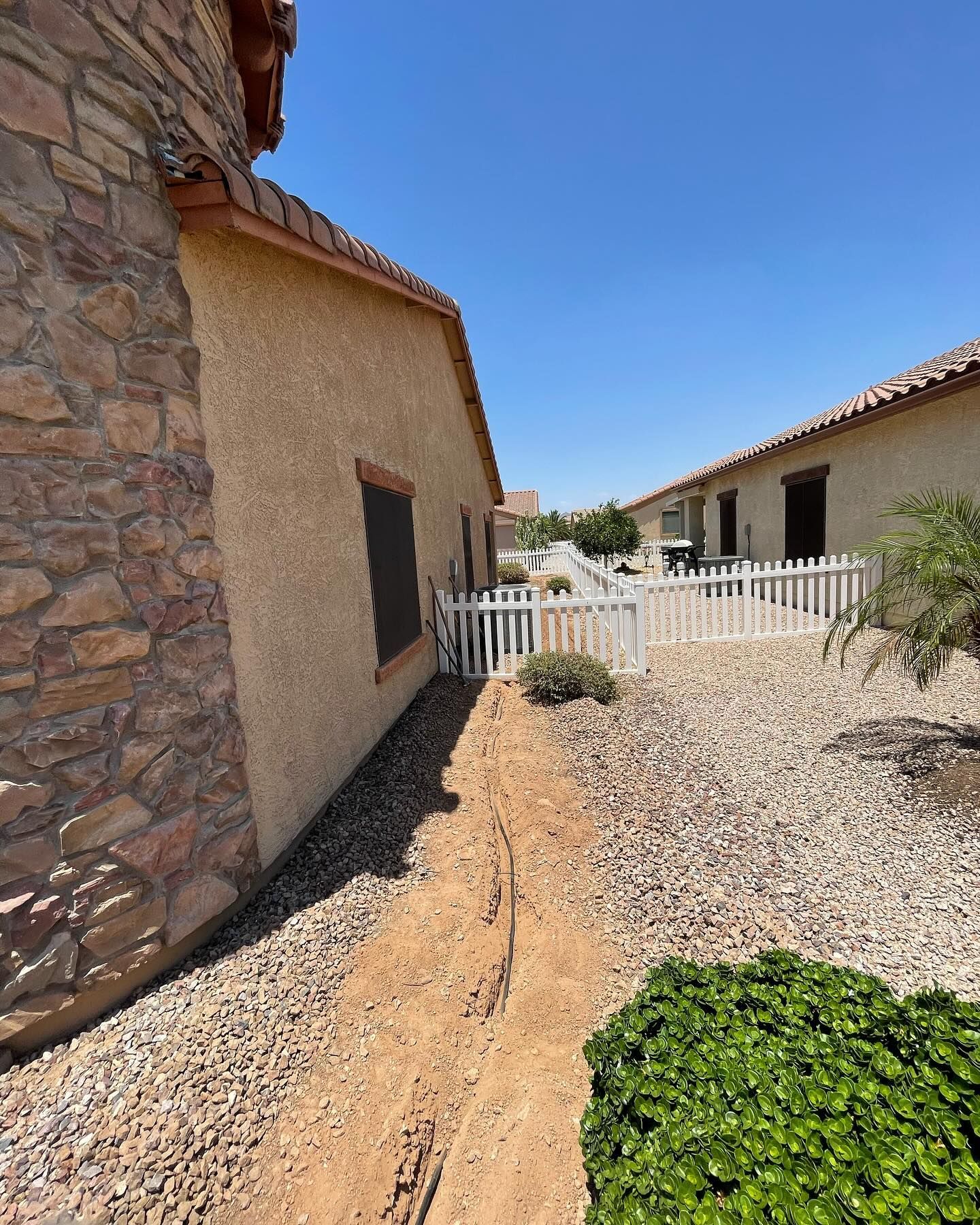 A narrow dirt trench alongside a building with a stone facade, small white picket fence, and gravel landscaping.