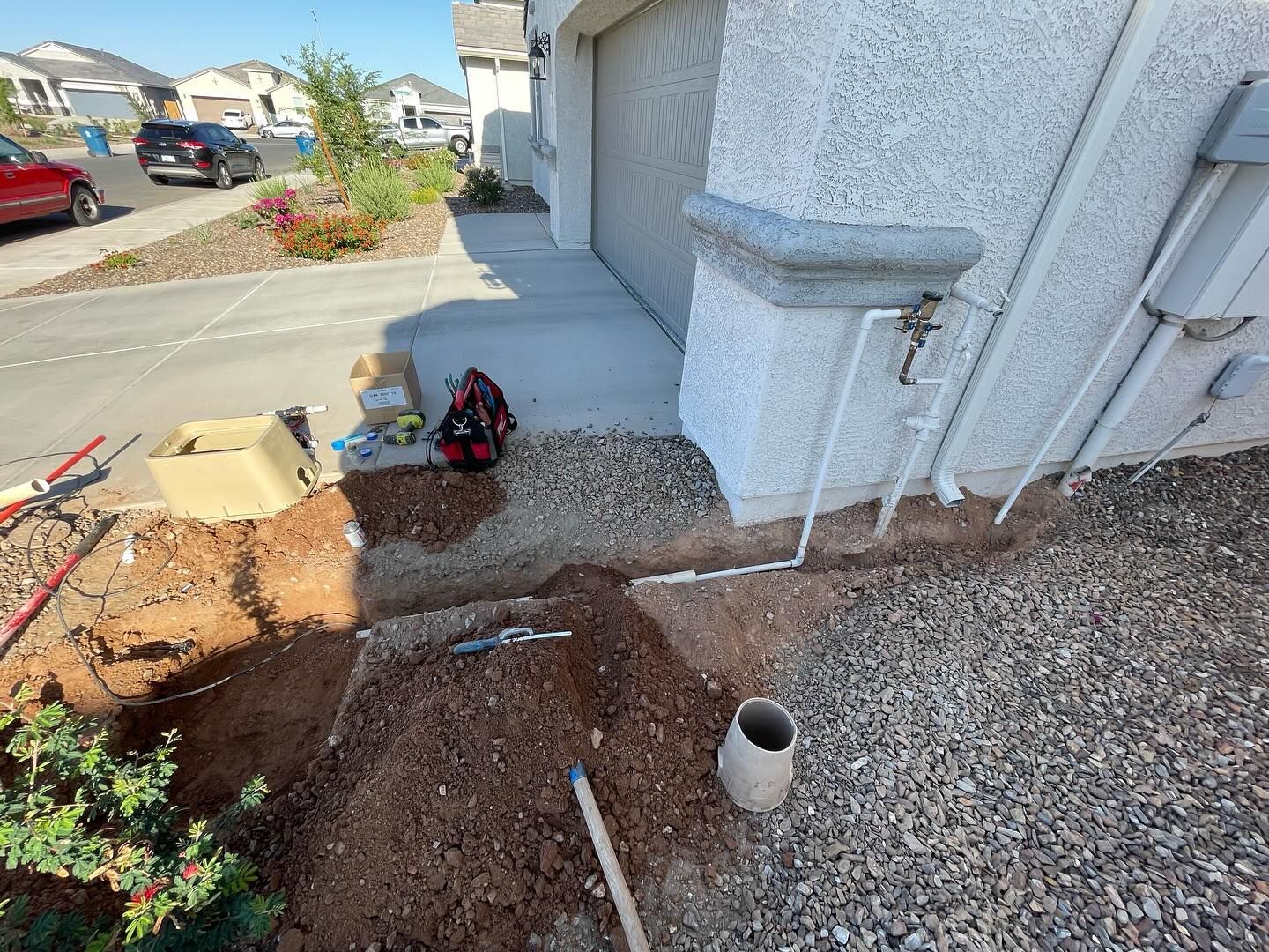 A dug-up yard area with gravel, tools, and a garage. A house is in the background on a sunny day.