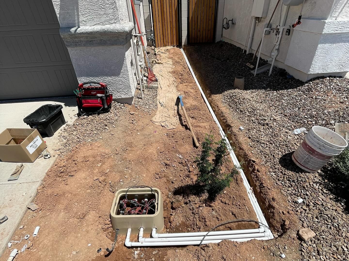 Trench dug along a pathway with irrigation components, boxes, and tools in a residential area.
