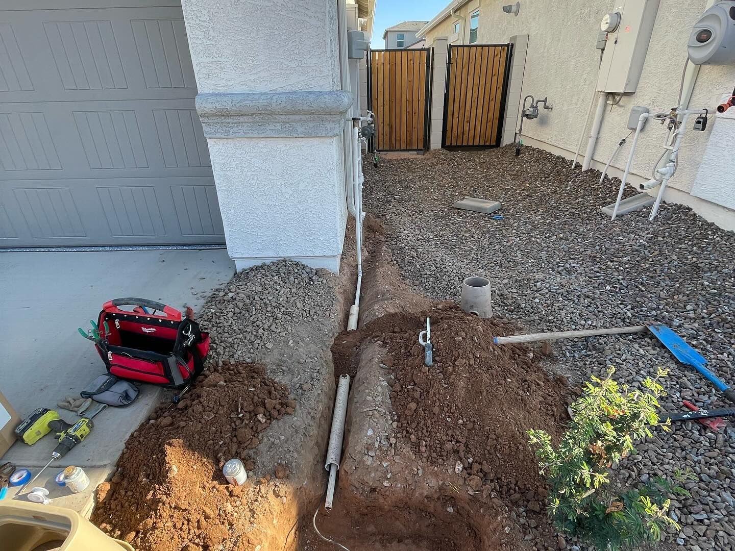 Trench with pipes along a house, construction tools, gravel and dirt, leading to a wooden gate.