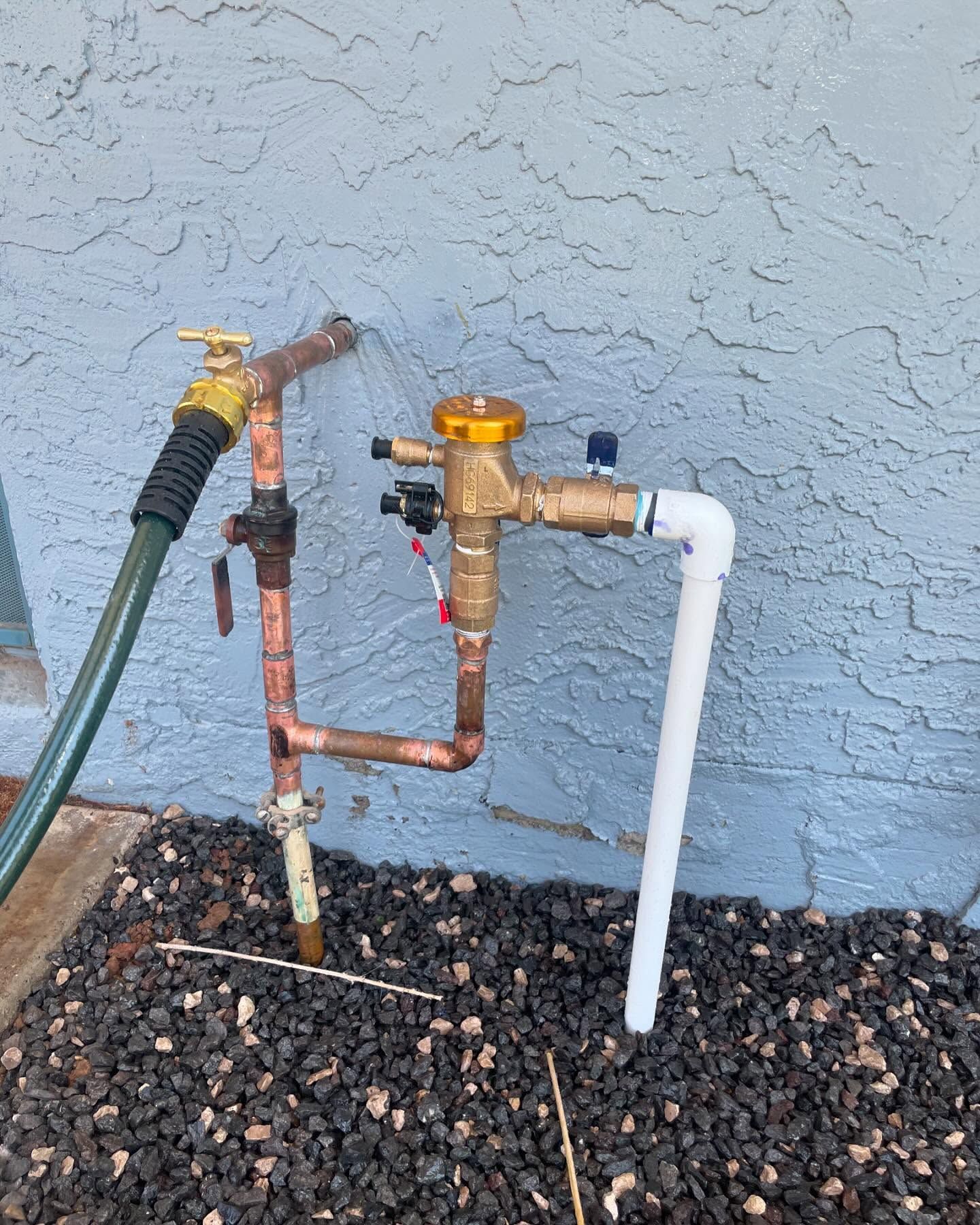 Irrigation system with a backflow preventer, pipes, and a hose nozzle against a stucco wall.