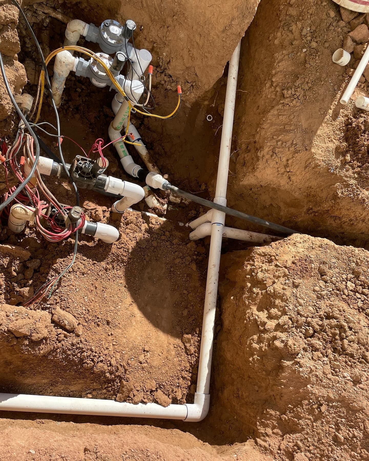 Irrigation system components in an excavated trench. White PVC pipes, valves, and wiring in brown soil.