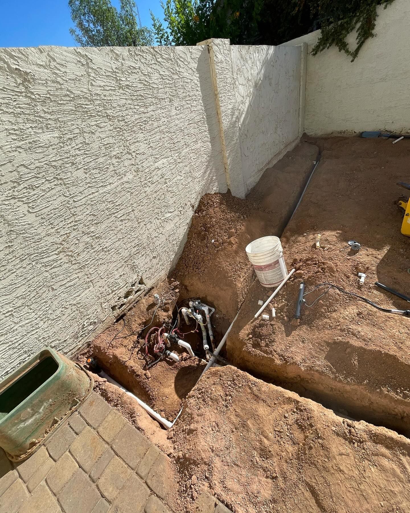 Irrigation system being installed; trenches in dirt, pipes and valves near a textured wall and a green box.
