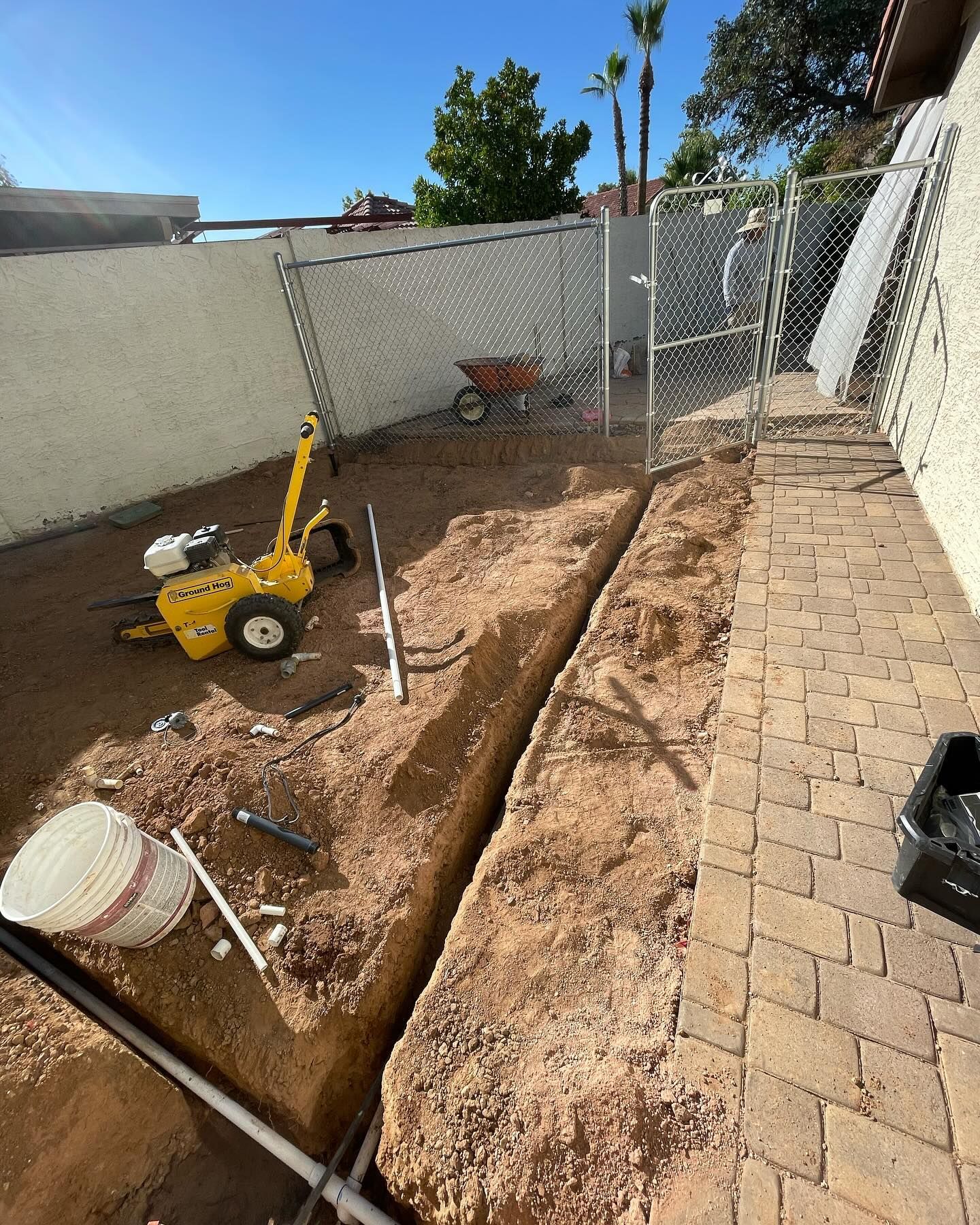 A trench dug in a backyard for plumbing. Yellow trencher and materials nearby.