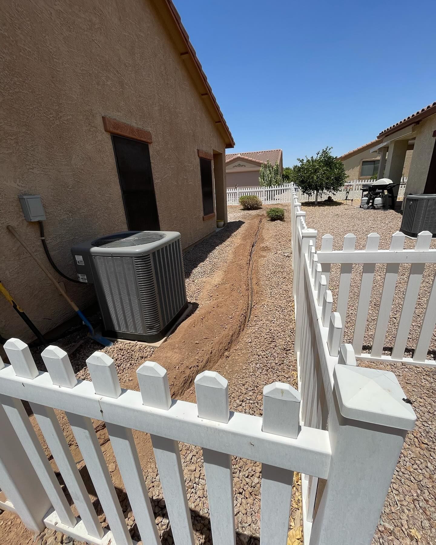 Trench dug along a house foundation, between a white picket fence and AC unit, under a clear blue sky.
