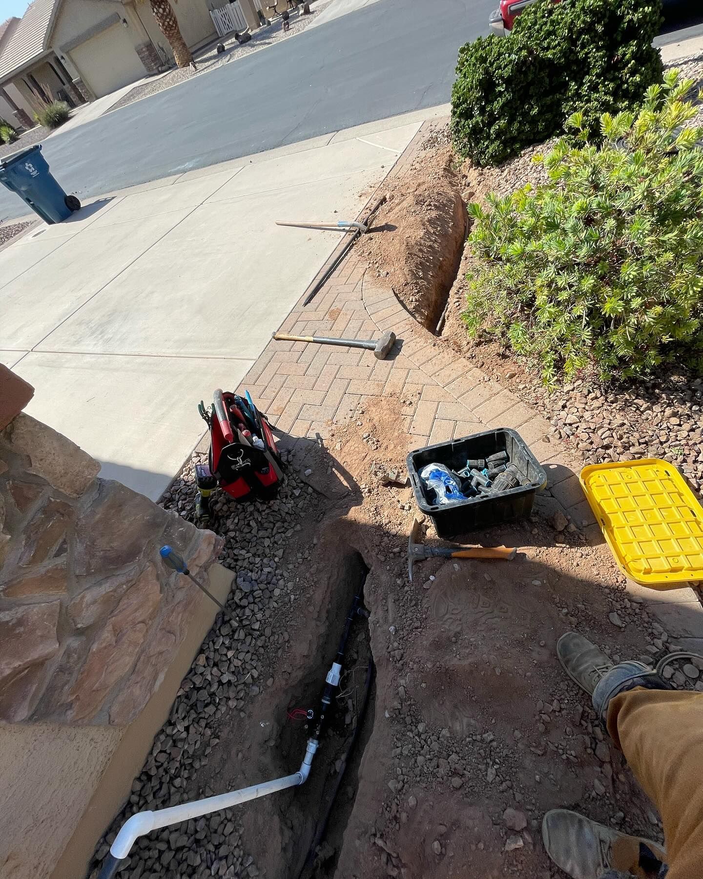 Trench dug along a driveway, plumbing work in progress. Tools and materials are visible, with a sidewalk, street, and plants.