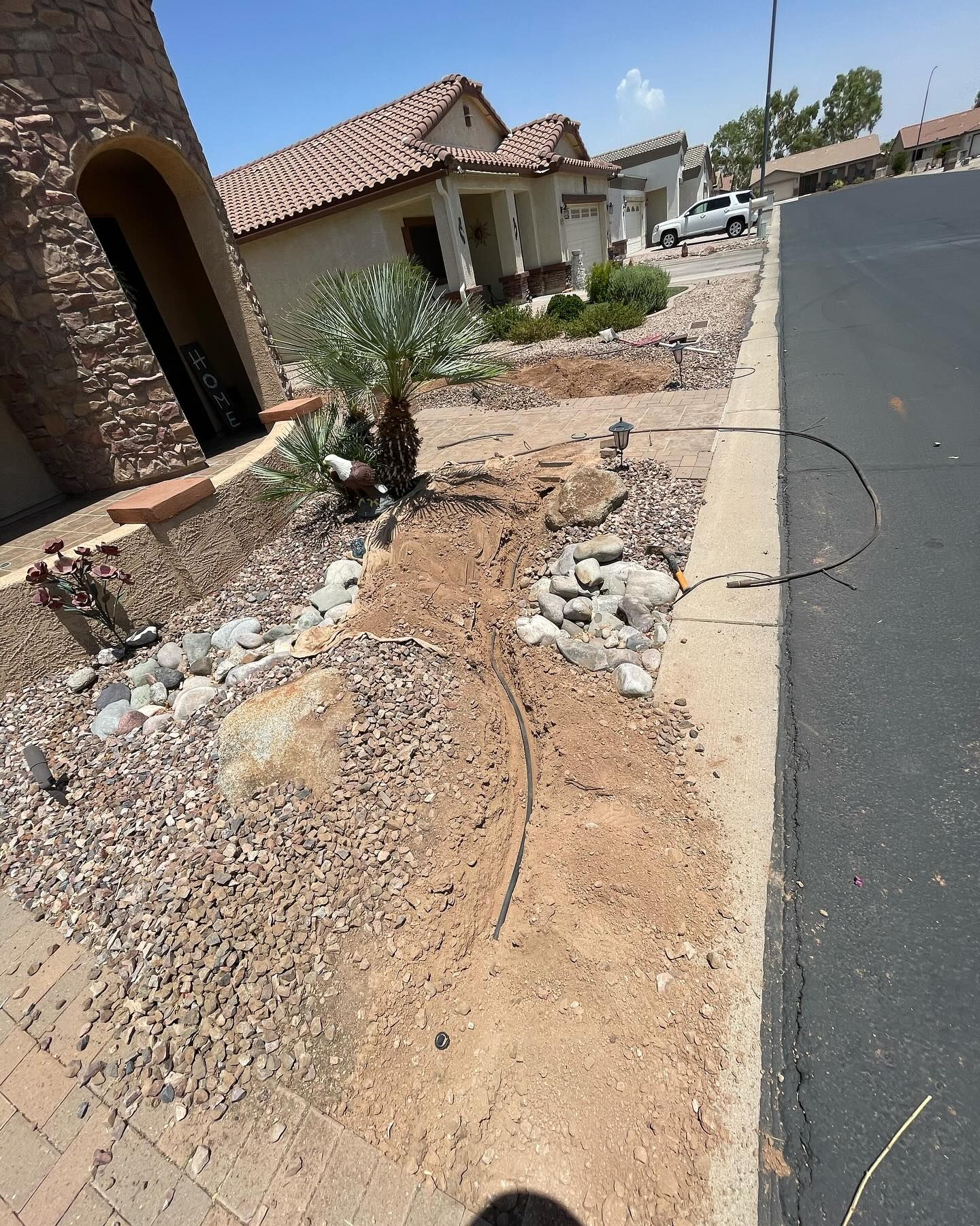 A sunny shot shows a house's dirt landscaping bordering a street. A palm tree, rocks, and irrigation tubing are visible.