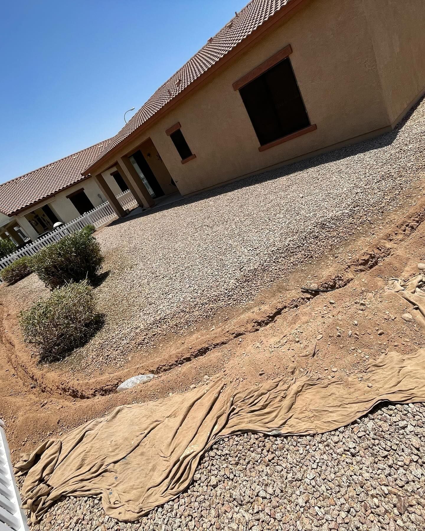 Gravel yard with a tan house and brown roof; dry plants, dirt, and a tan fabric border.