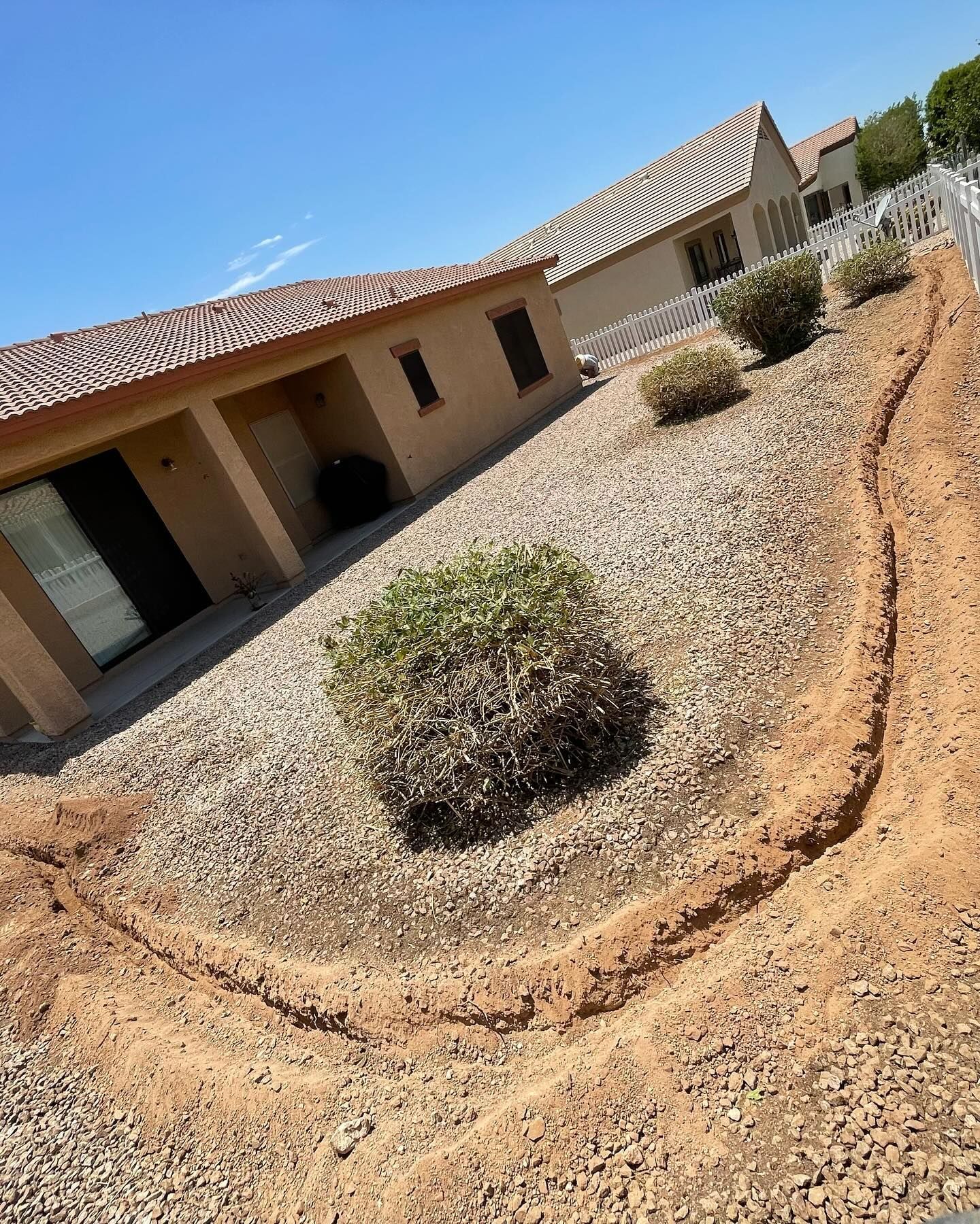 A trench being dug in a yard filled with gravel next to a beige house under a blue sky.