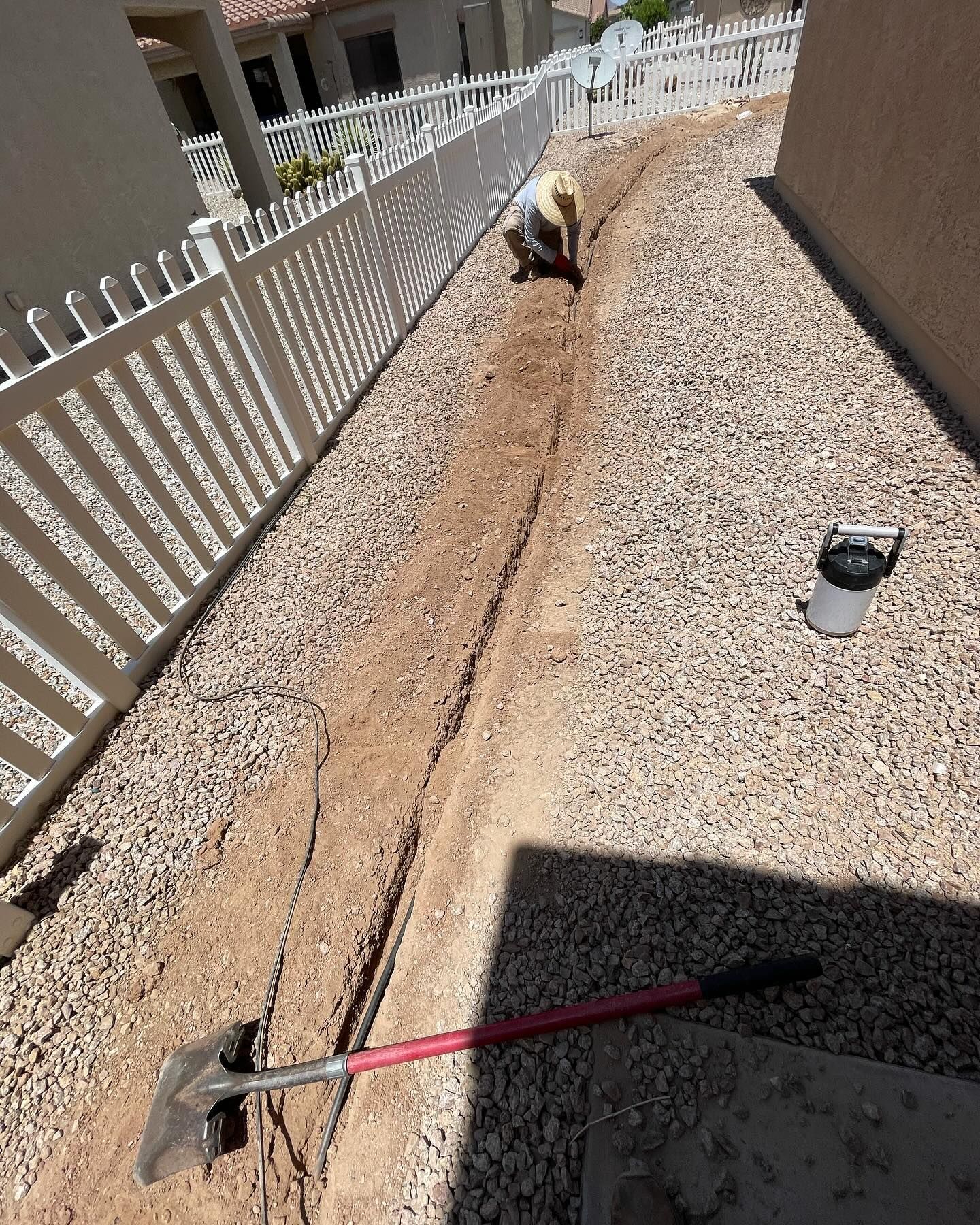 A person digging a trench in a gravel-covered yard next to a white picket fence and a building.