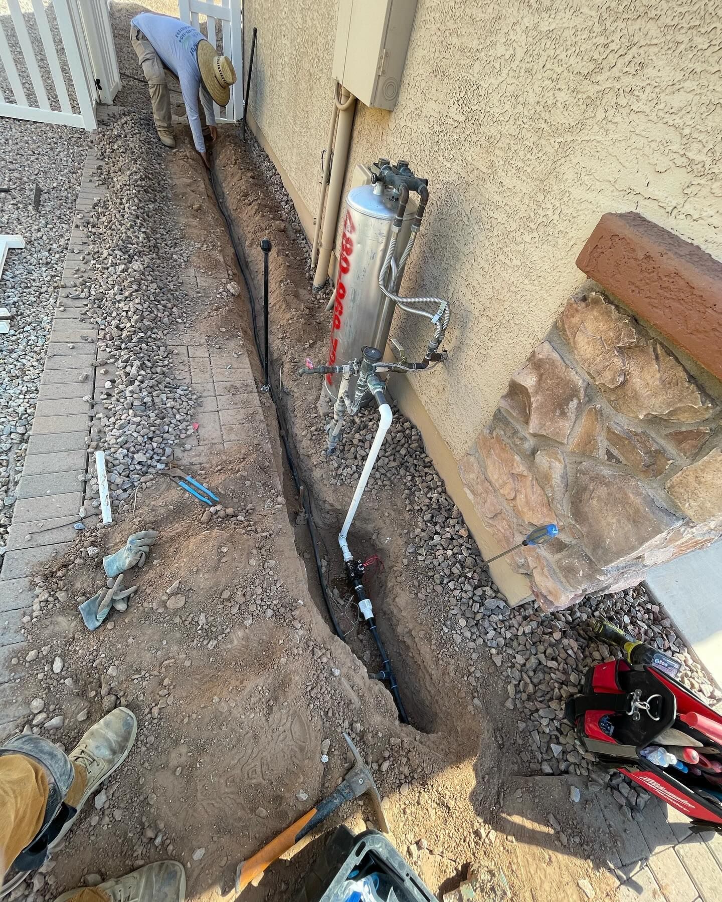 Man working on a buried pipe trench beside a building. Dirt, rocks, tools, and plumbing components are visible.