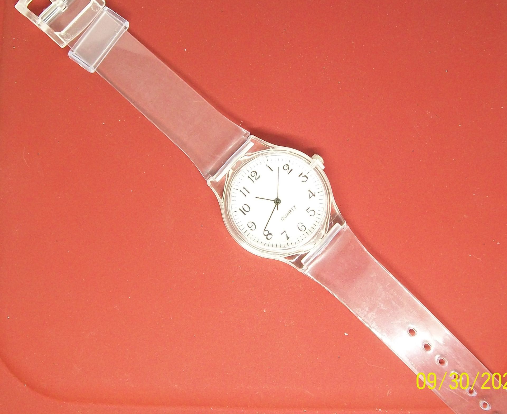 Een helder, rond horloge met een witte wijzerplaat op een rode ondergrond. Daaronder staat een label: 