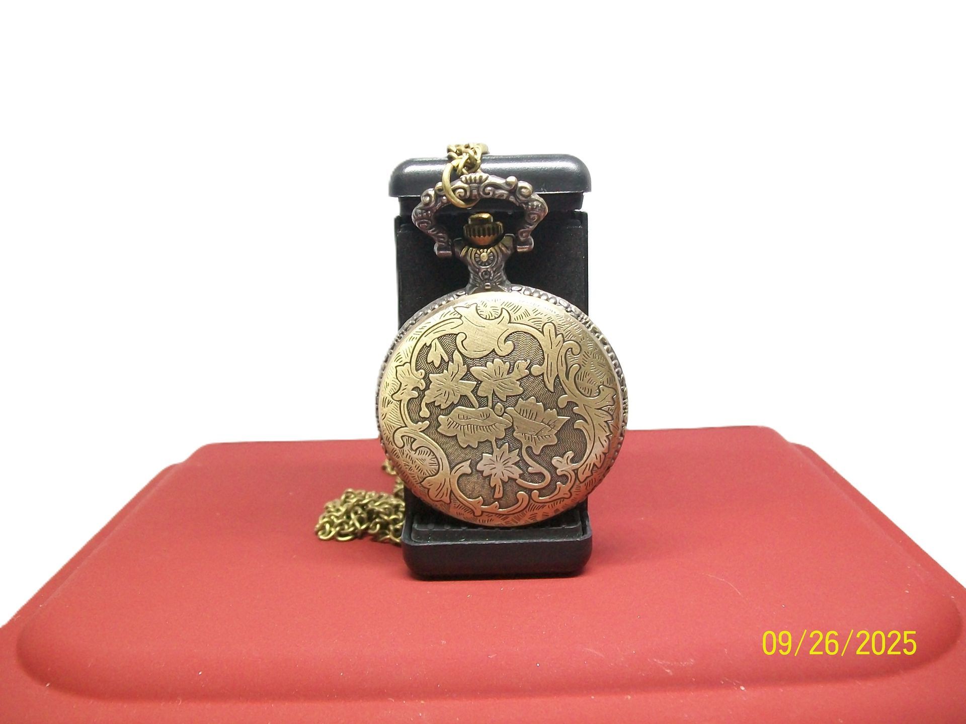 Zakhorloge op een rood oppervlak, met een gouden ketting en gedeeltelijk open deksel, daarachter een zwarte kast, datum zichtbaar: 26-09-2005.