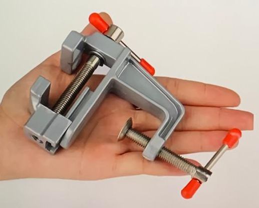 Een kleine zilveren bankschroef met rode handvatten, vastgeklemd aan de rand van een houten oppervlak.