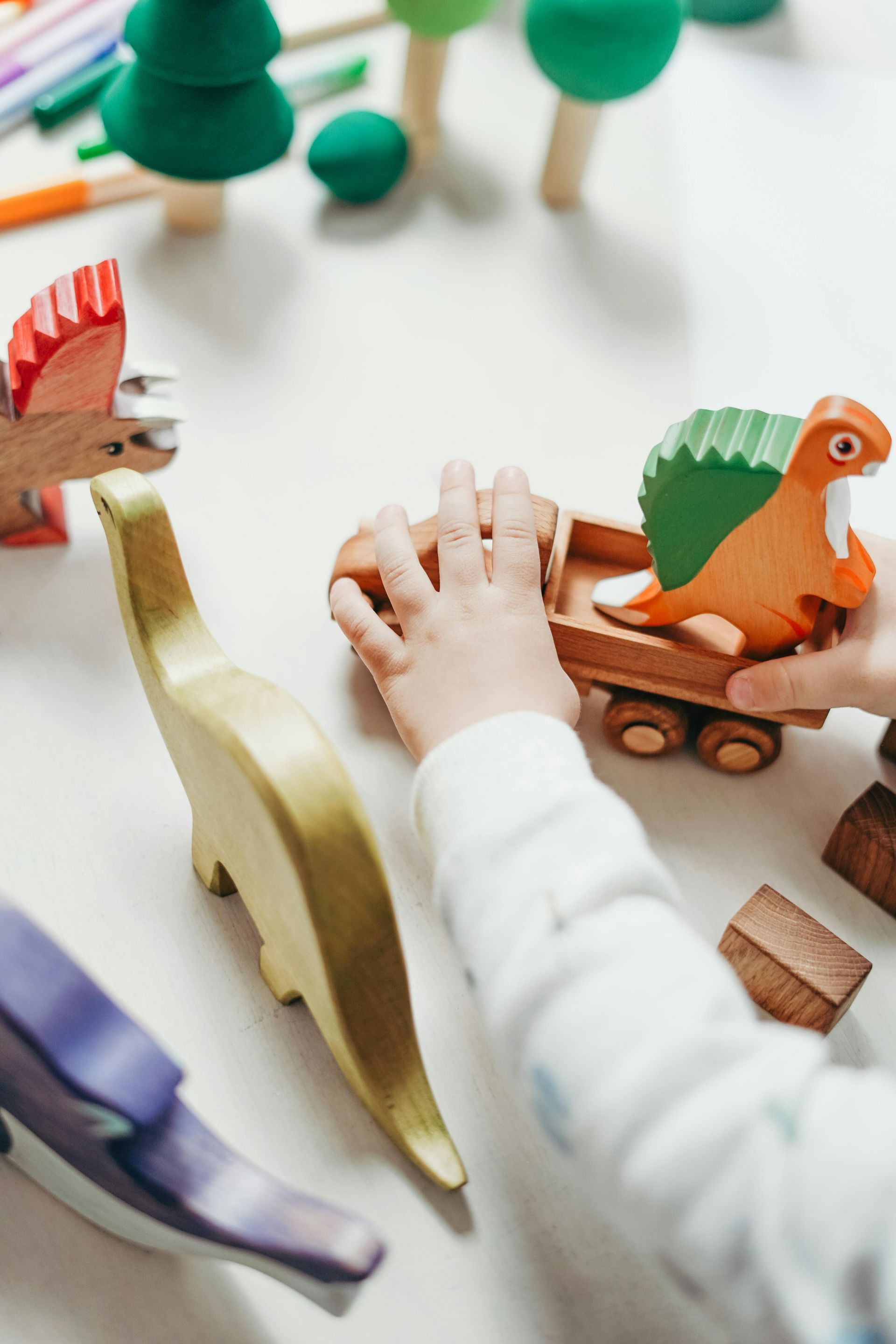 Een kind speelt met houten dinosaurussen en een vrachtwagen op een wit oppervlak, omringd door houten bomen.