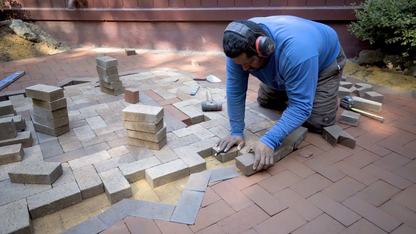 Man laying brick pavers on a patio with ear protection. Brick stacks and dirt visible.