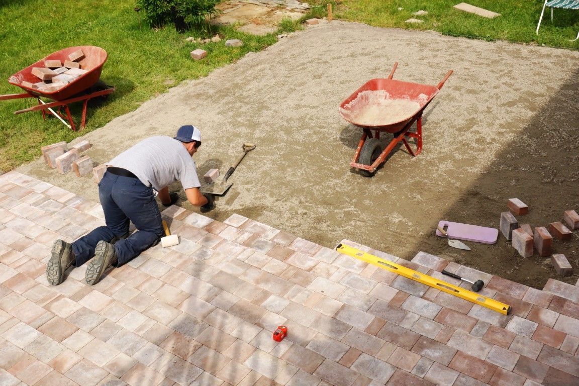 Person laying brick pavers on a patio with wheelbarrows, tools, and materials.