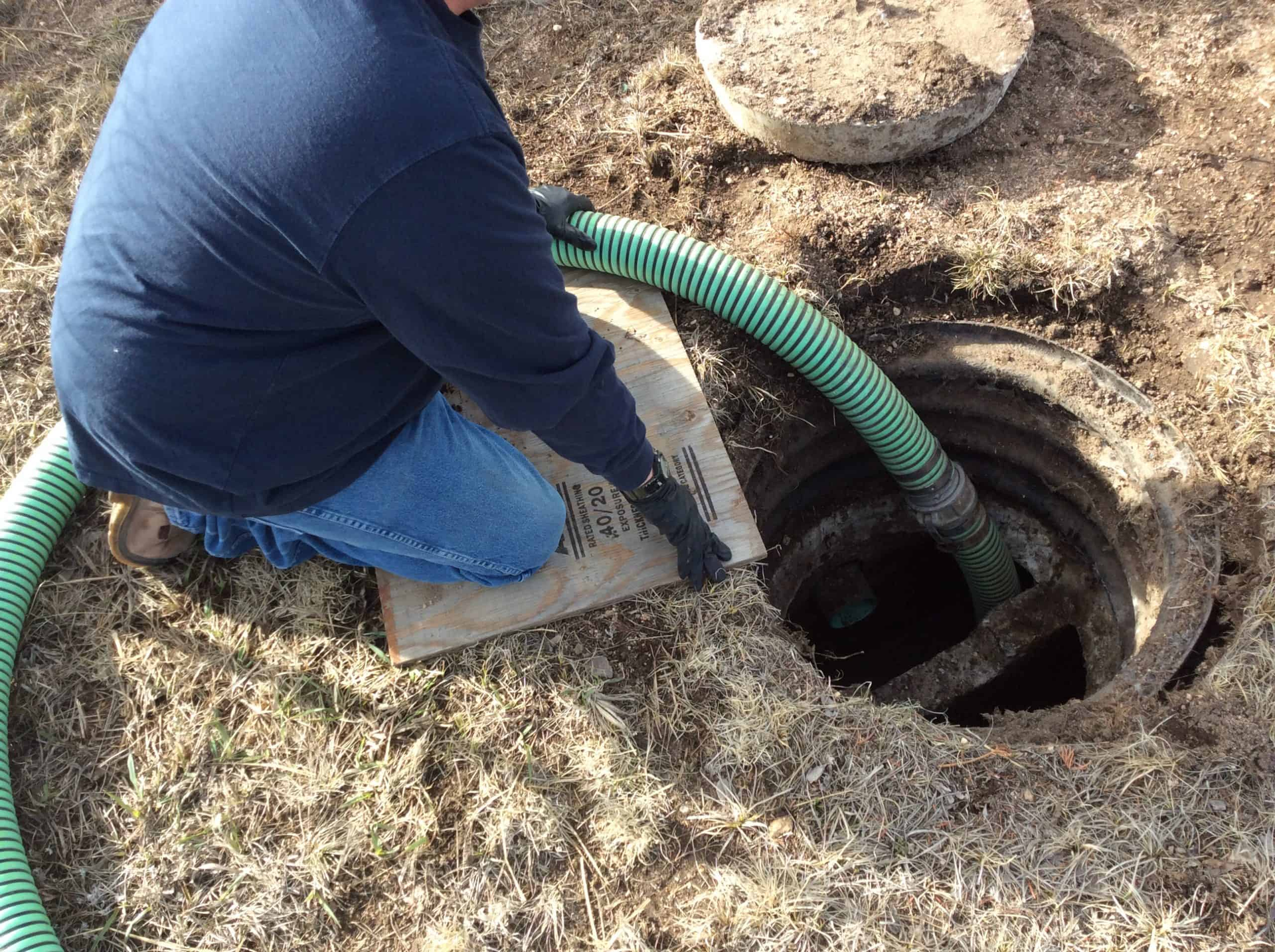 Person in blue jacket pumping a septic tank from an open access. Green hose, outdoor setting.