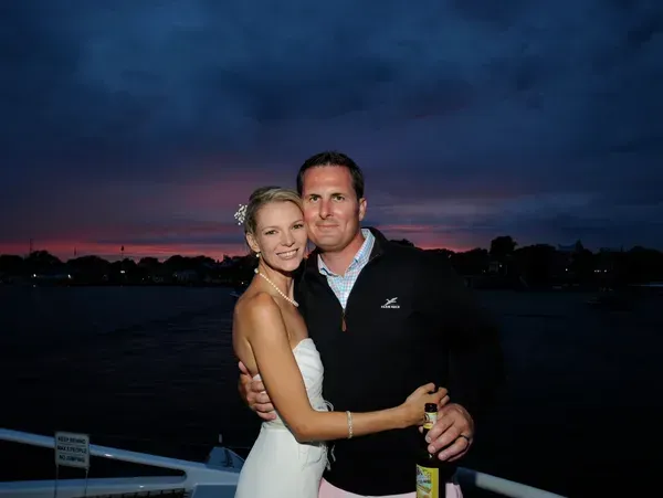 A couple embraces on a boat deck at twilight, with the woman in a white dress and the man in a dark zip-up jacket.
