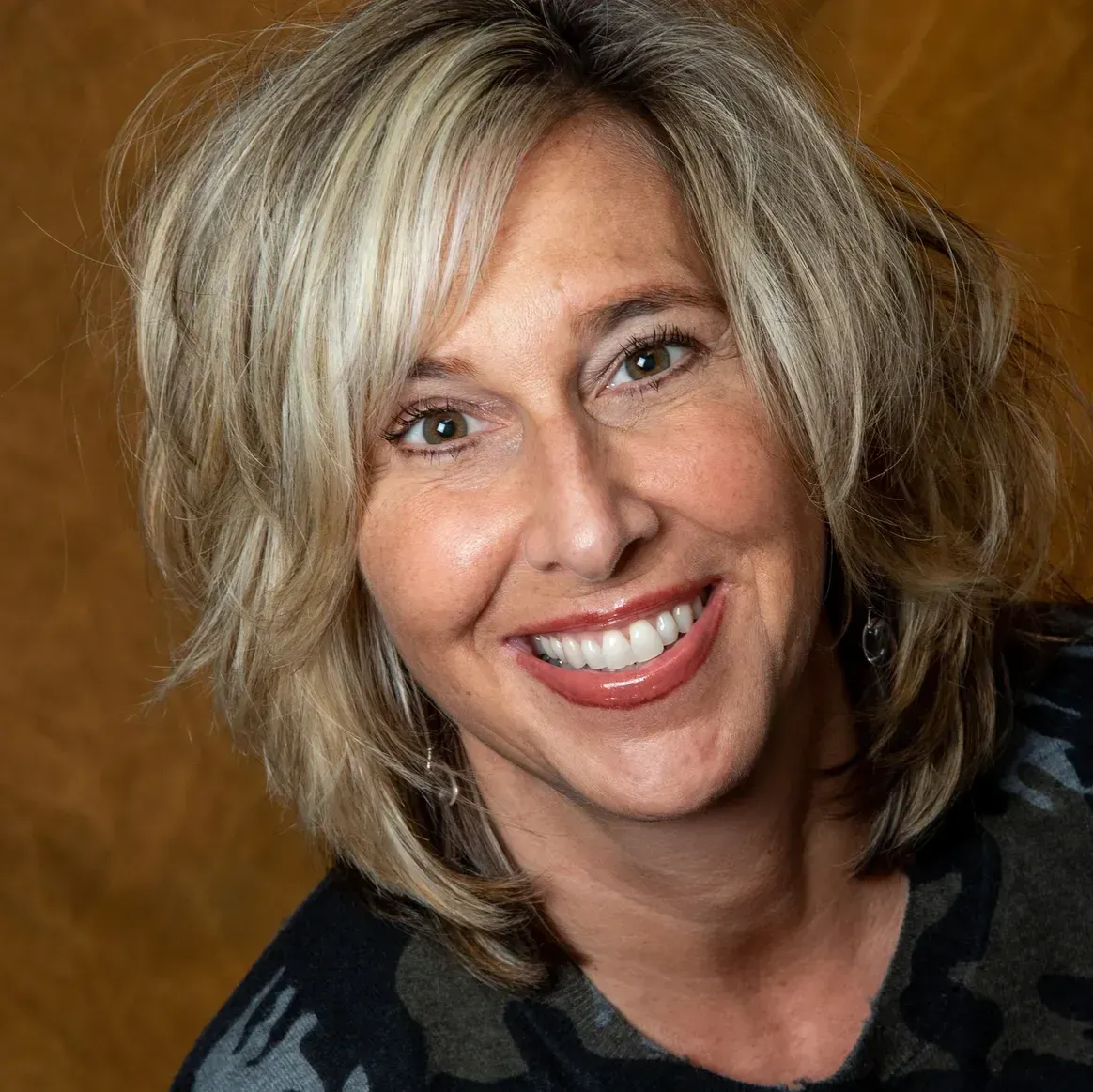 A smiling person with shoulder-length blonde hair, wearing a patterned dark top, against a mottled brown background.