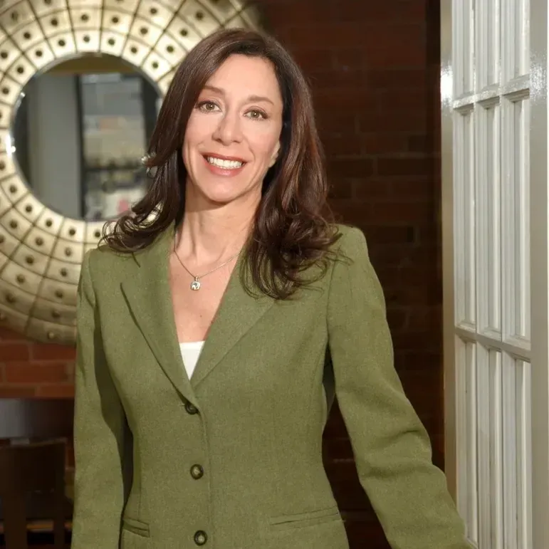 A professional person in a green blazer smiles in an indoor room with a round decorative mirror and white door frame.