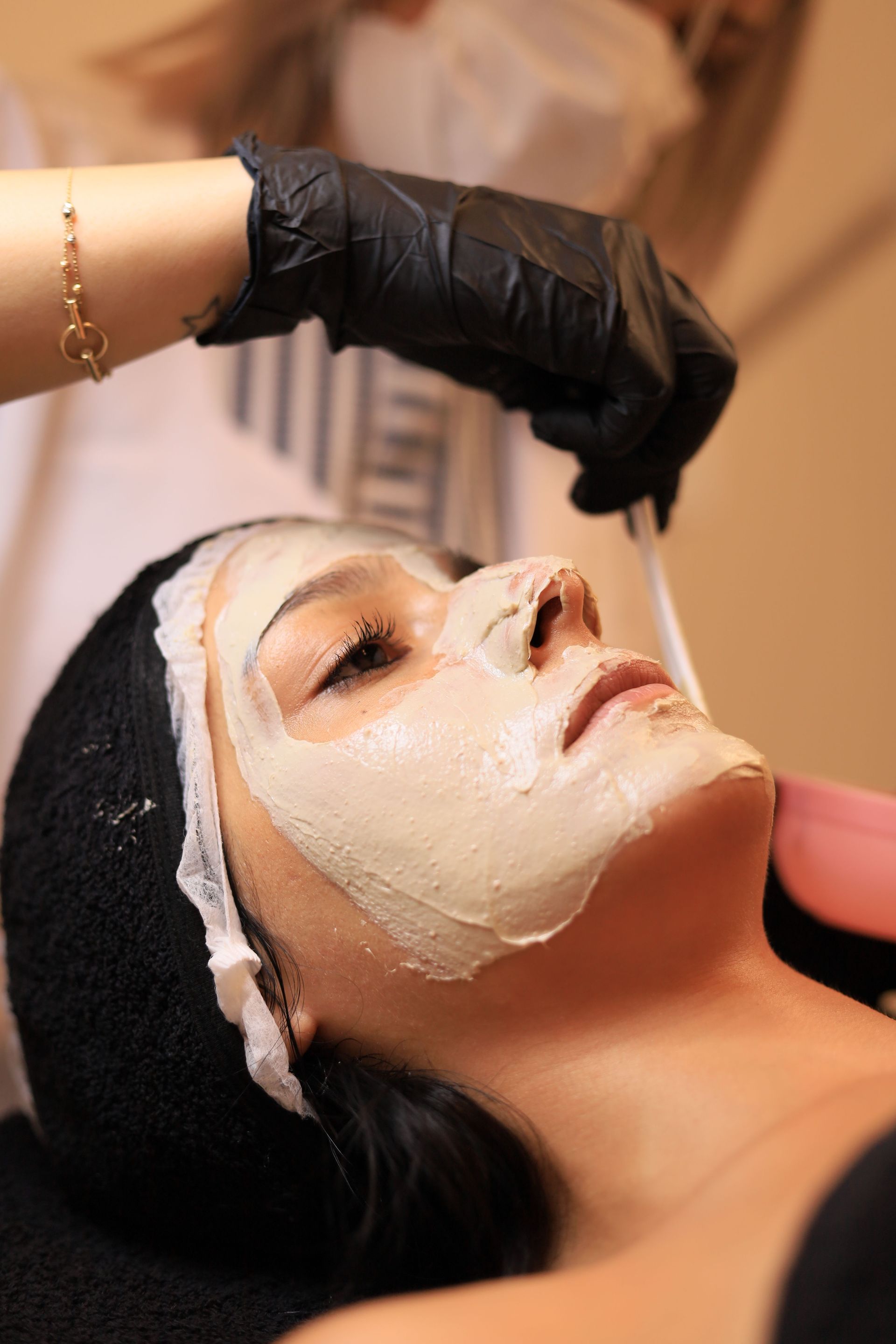 Facial mask application at a spa, with a gloved hand smoothing cream over a reclining client’s face