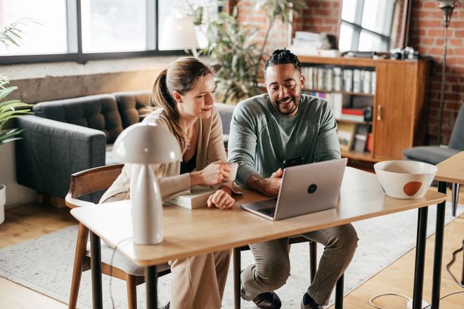 Two people looking at laptop screen at a table in a home office.