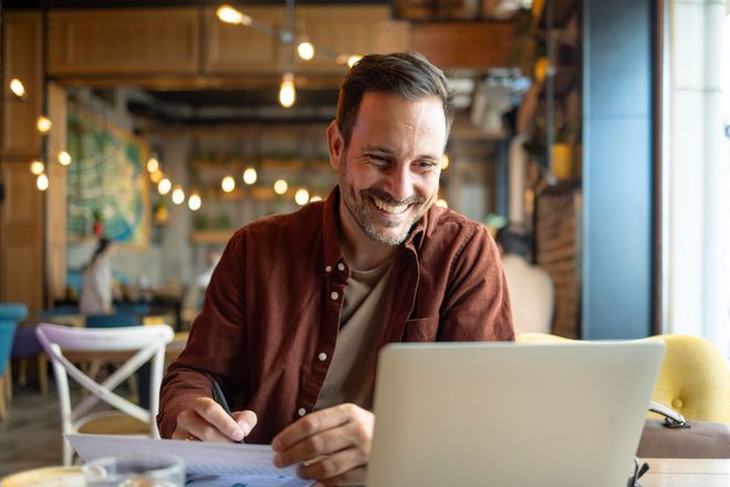 Man in brown shirt smiles while working on laptop and papers at a cafe.