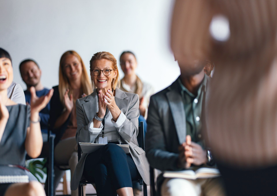Audience applauds a speaker during a presentation. People sit in chairs; some clap, smile, and laugh.