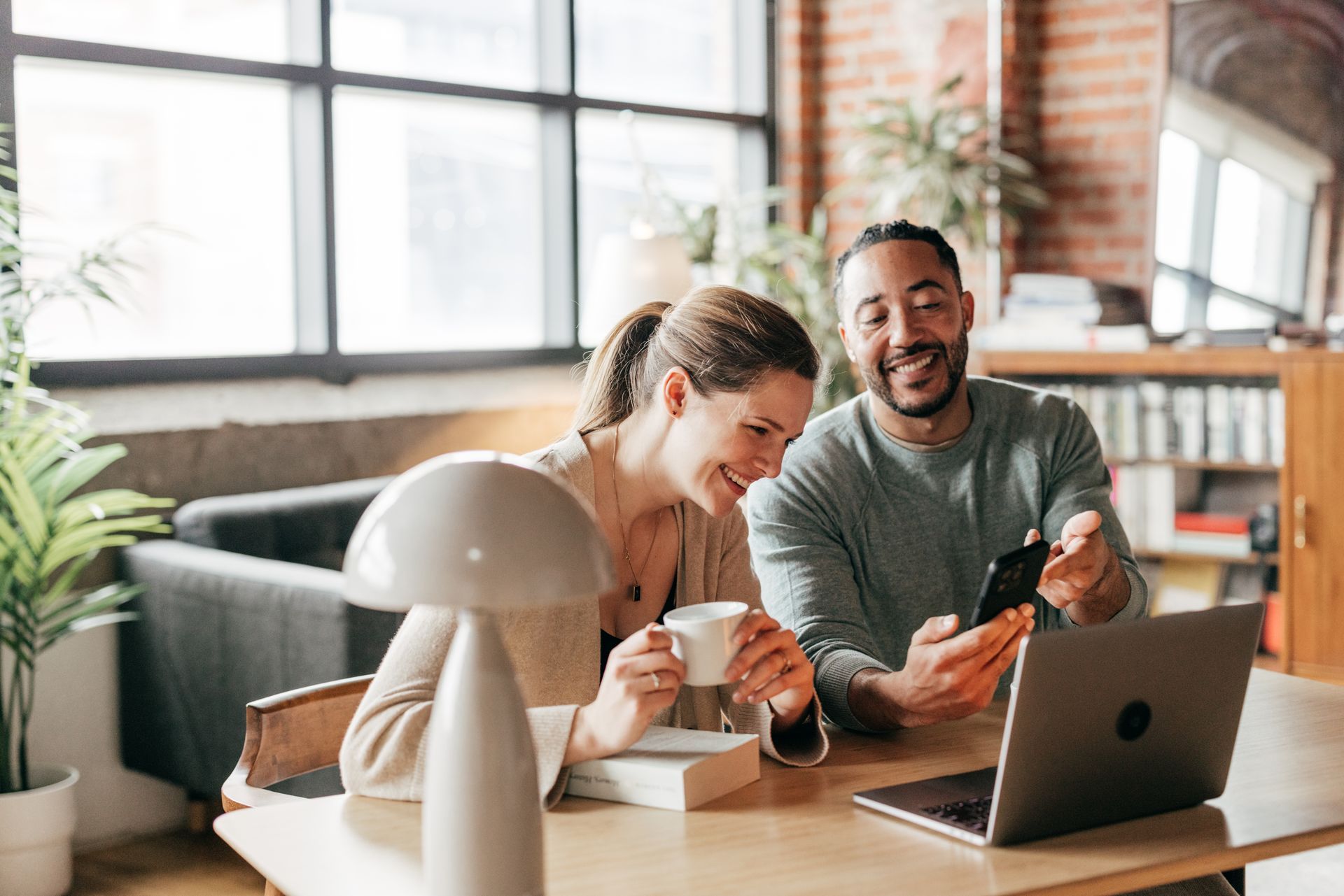 Couple smiles while looking at a smartphone, laptop open on table. Indoor setting, brick wall, lamp and plants visible.
