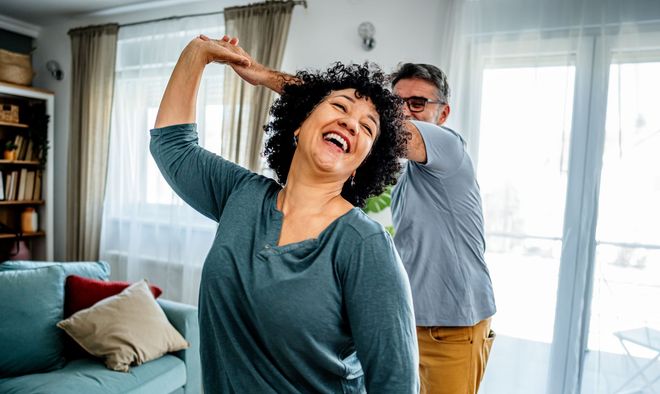 Couple dancing indoors; woman laughs with arm raised, man holds her hand. Bright room, natural light.