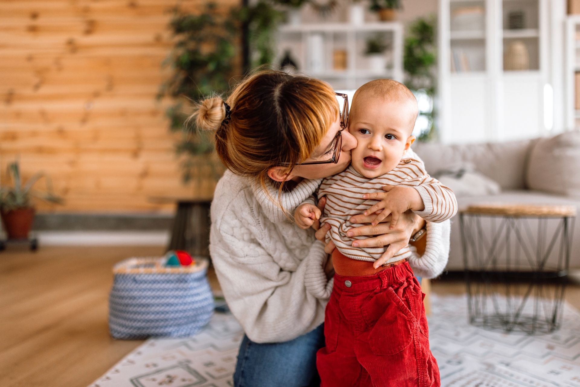 Woman kisses a baby held in her arms; both are smiling in a cozy living room.