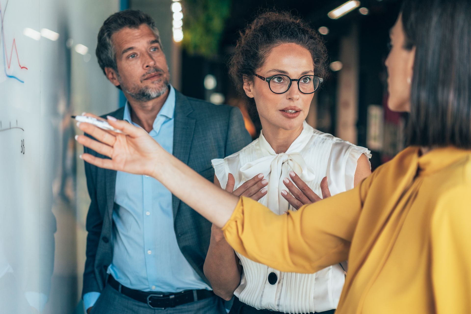 Three people in a modern office, one pointing at whiteboard, two others looking on attentively.