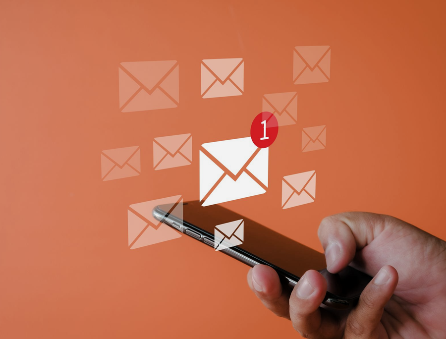Hand holding a phone with an email icon and a notification bubble, surrounded by email envelope icons on an orange background.