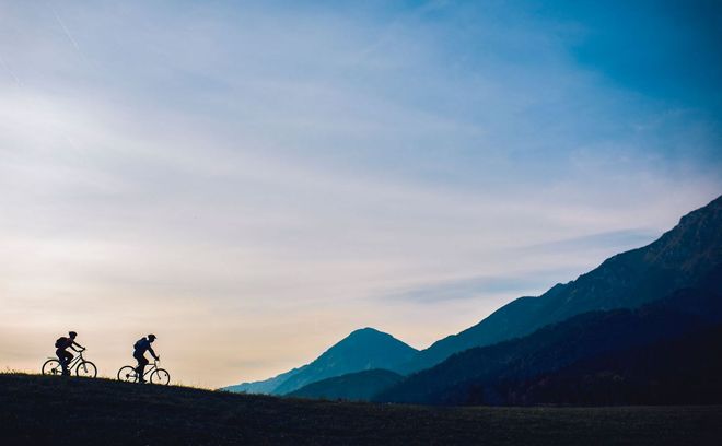 Two cyclists silhouetted on a hillside, riding towards a mountain range under a cloudy sky.