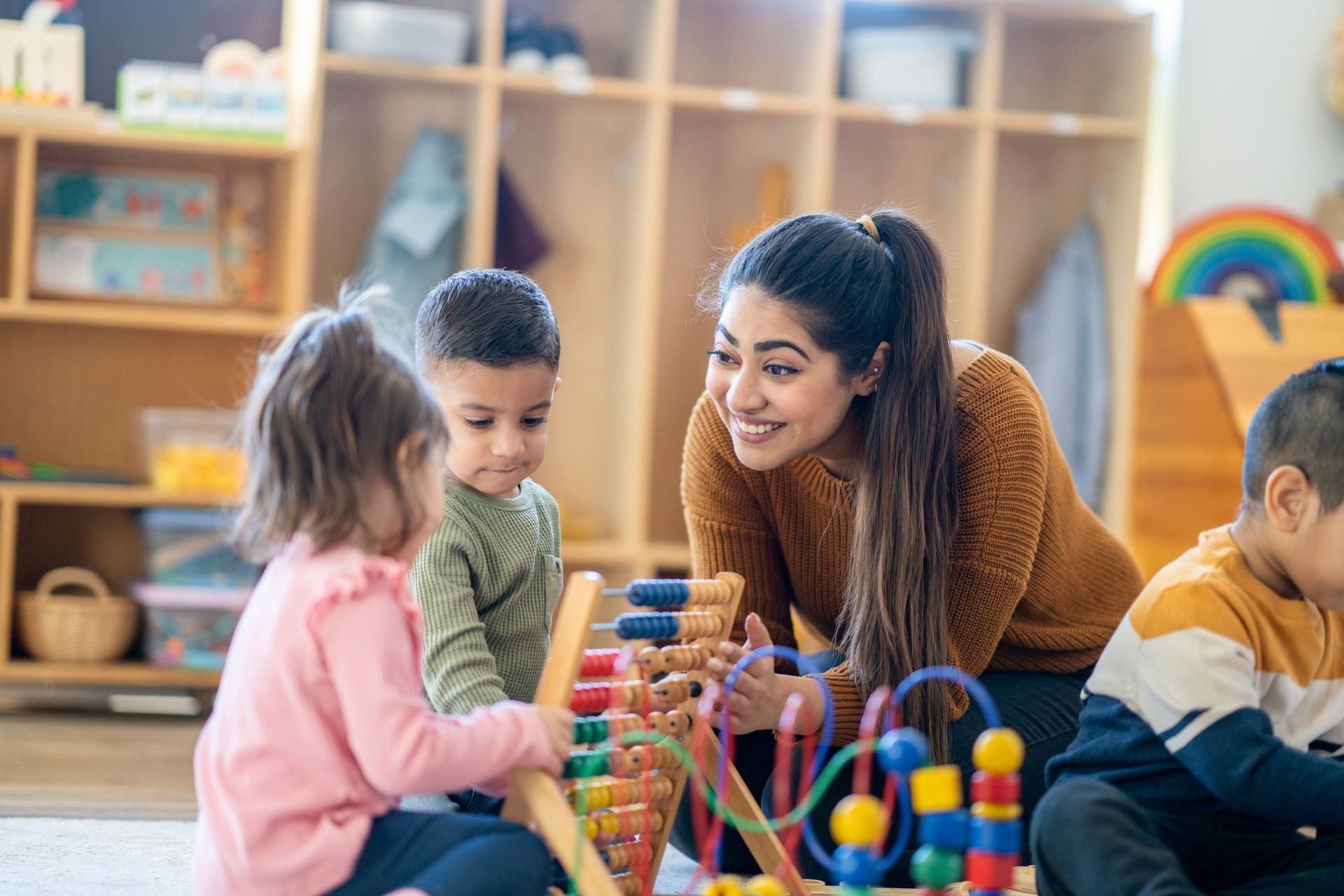 A pre-kindergarten teacher is seated on the floor with students as they play with various toys.
