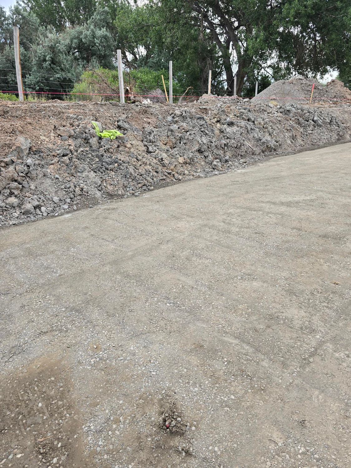 A gravel road borders a dark soil pile. Trees and fence posts are in the background.
