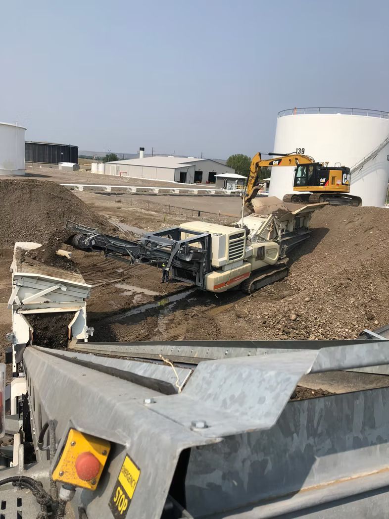 Heavy machinery sorting dirt at a construction site near large white tanks on a sunny day.