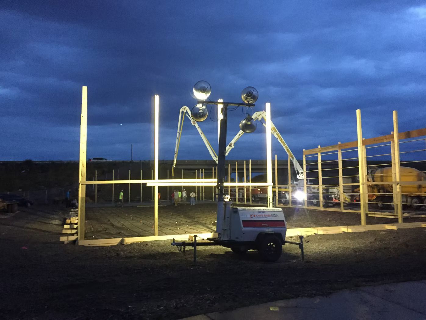 Construction site at dusk, illuminated by a mobile light tower. Wood framing is in progress.