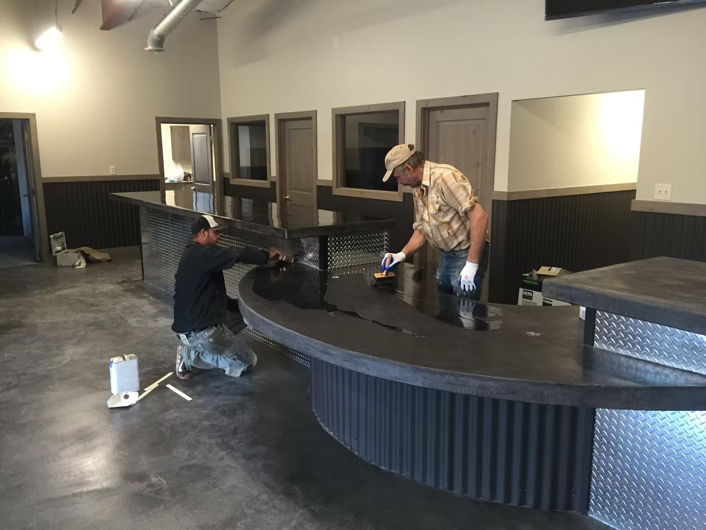 Two workers refinish a dark bar counter with corrugated metal accents. Indoor setting.