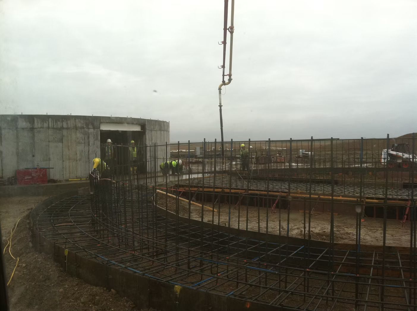 Construction site with rebar framework, concrete pouring from a boom, workers in vests.