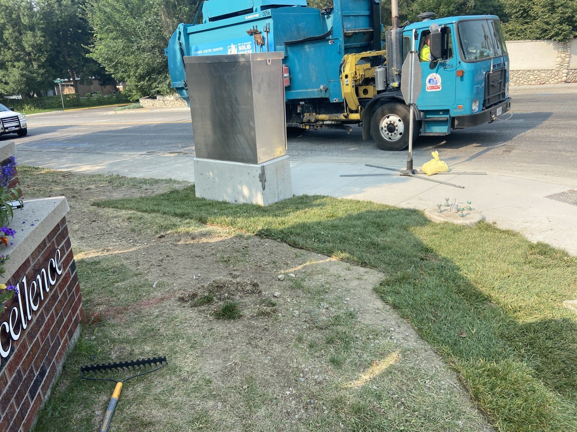 Garbage truck parked next to a dark gray box on a grassy area, near a brick building with a sign.